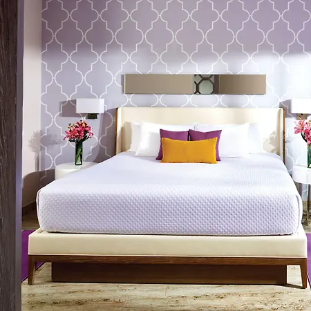 The Fives Beach Hotel & Residences - All Senses Inclusive Playa del Carmen Beds photo The photo shows a well-lit hotel bedroom with a focus on a comfortable queen-sized bed. The bed has a white quilted coverlet and is adorned with several pillows: two white standard pillows, two smaller square pillows in a deep purple, and a rectangul