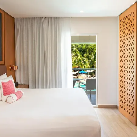 The Fives Beach Hotel & Residences - All Senses Inclusive Playa del Carmen Exterior photo The photo shows a well-lit hotel bedroom with a focus on the bed and a glimpse of an outdoor space. 

In the foreground is a large bed with white linens, several pillows including two fluffy white ones and a decorative pink bolster pillow. Two rolled