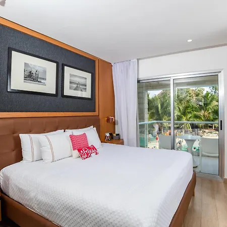 The Fives Beach Hotel & Residences - All Senses Inclusive Playa del Carmen Exterior photo The photo shows a well-lit hotel bedroom with a large bed as the focal point. The bed has a white quilted comforter, several pillows including white and striped ones, and a decorative pink pillow with white polka dots. The headboard is a brown leathe