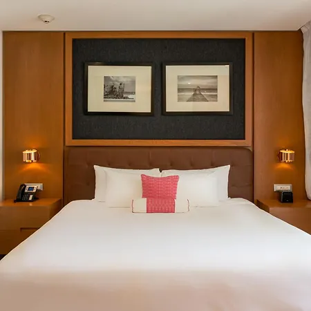 The Fives Beach Hotel & Residences - All Senses Inclusive Playa del Carmen Exterior photo The photo shows a well-lit hotel bedroom featuring a comfortable queen-sized bed as the focal point. The bed is neatly made with crisp white linens, several white pillows, and a decorative stack of folded pink towels. A small, square pink pillow adds