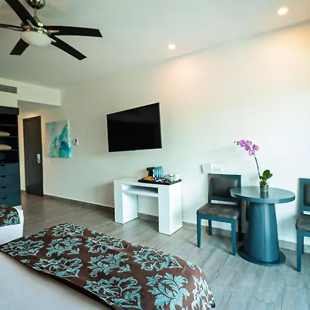 The Fives Beach Hotel & Residences - All Senses Inclusive Playa del Carmen Exterior photo The photo shows a modern hotel room with a clean and minimalist design. The walls are painted a bright white, and the flooring is a light gray wood-look laminate. 

In the foreground, a bed with a patterned brown and teal bedspread is partially visib