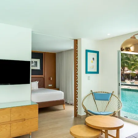 The Fives Beach Hotel & Residences - All Senses Inclusive Playa del Carmen Exterior photo The photo shows a bright and modern hotel room with a view of a beautiful swimming pool. 

The room is divided into a sleeping area and a living area. The sleeping area features a large bed with white linens and a wooden headboard. A flat-screen TV i