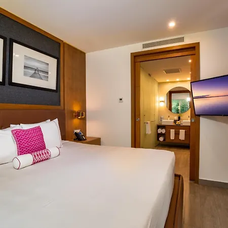 The Fives Beach Hotel & Residences - All Senses Inclusive Playa del Carmen Exterior photo The photo shows a modern hotel room with a comfortable king-sized bed as the central focus. The bed is neatly made with white linens, several pillows including two larger white ones with thin stripes and a vibrant pink decorative pillow, and a cylind